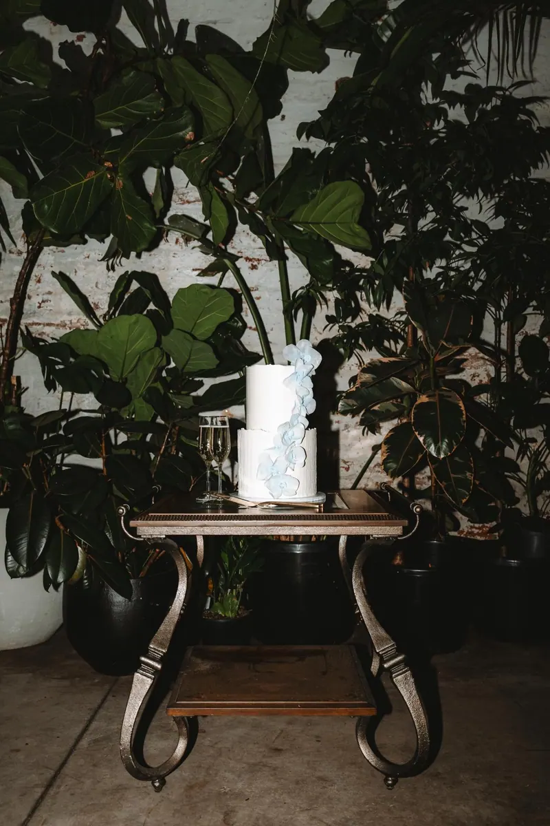 A floral wedding cake and champagne on an antique table, surrounded by lush greenery.